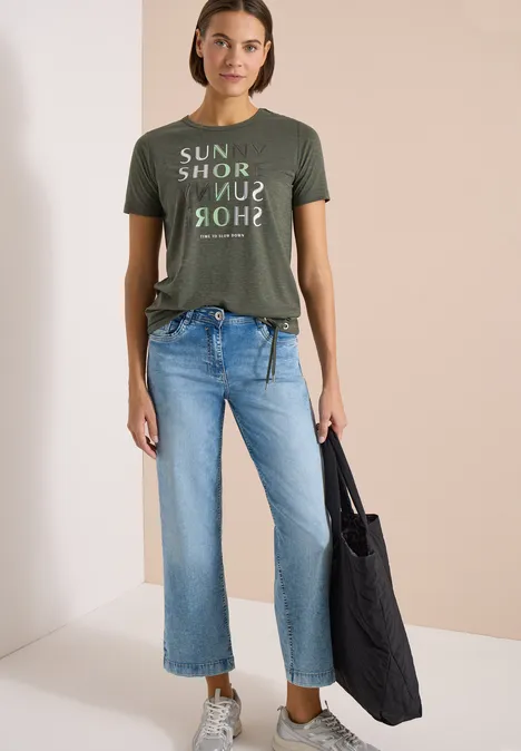 Frau im grünen T-Shirt mit Aufschrift, blauen Jeans und einer schwarzen Stofftasche.